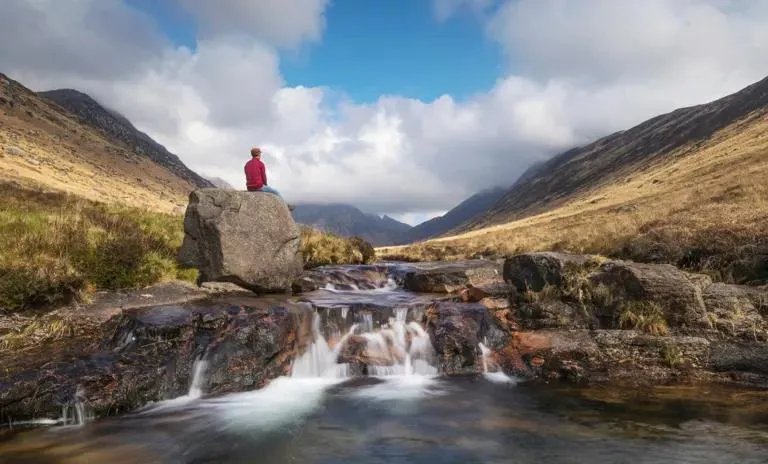 Person sits on top of a large boulder by a stream in an Arran Glen