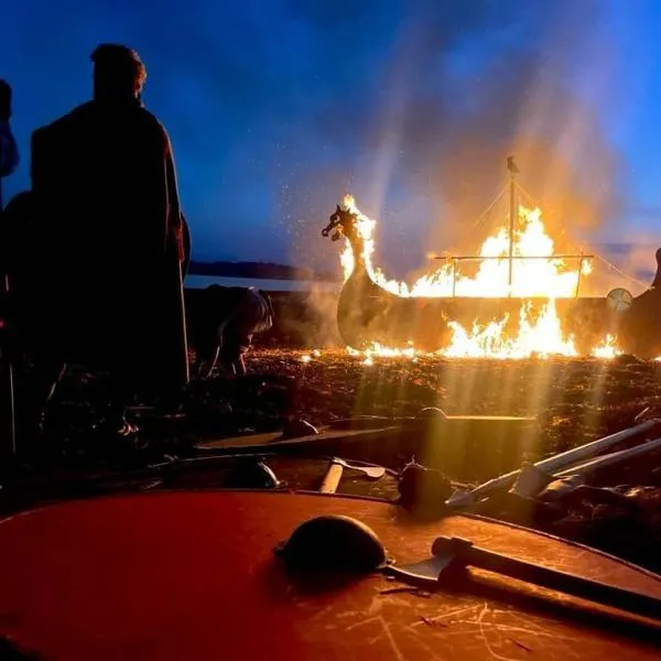 A burning longship on a beach with various items of viking paraphernalia strewn around about